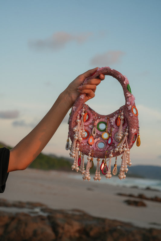 Dusty rose beaded Formentera bag with teardrop pendants and pearl fringe held against beach backdrop