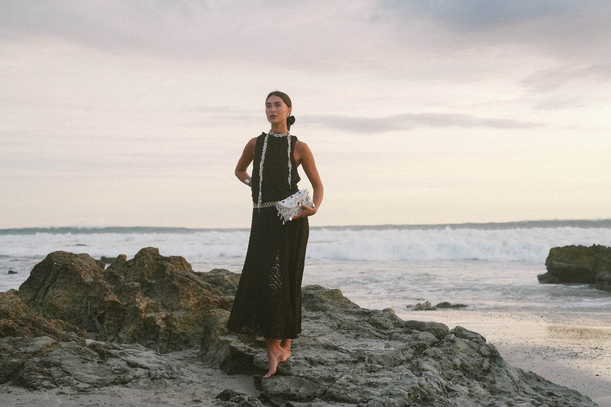 Woman in black sleeveless dress with beaded white bag standing on rocky beach at sunset